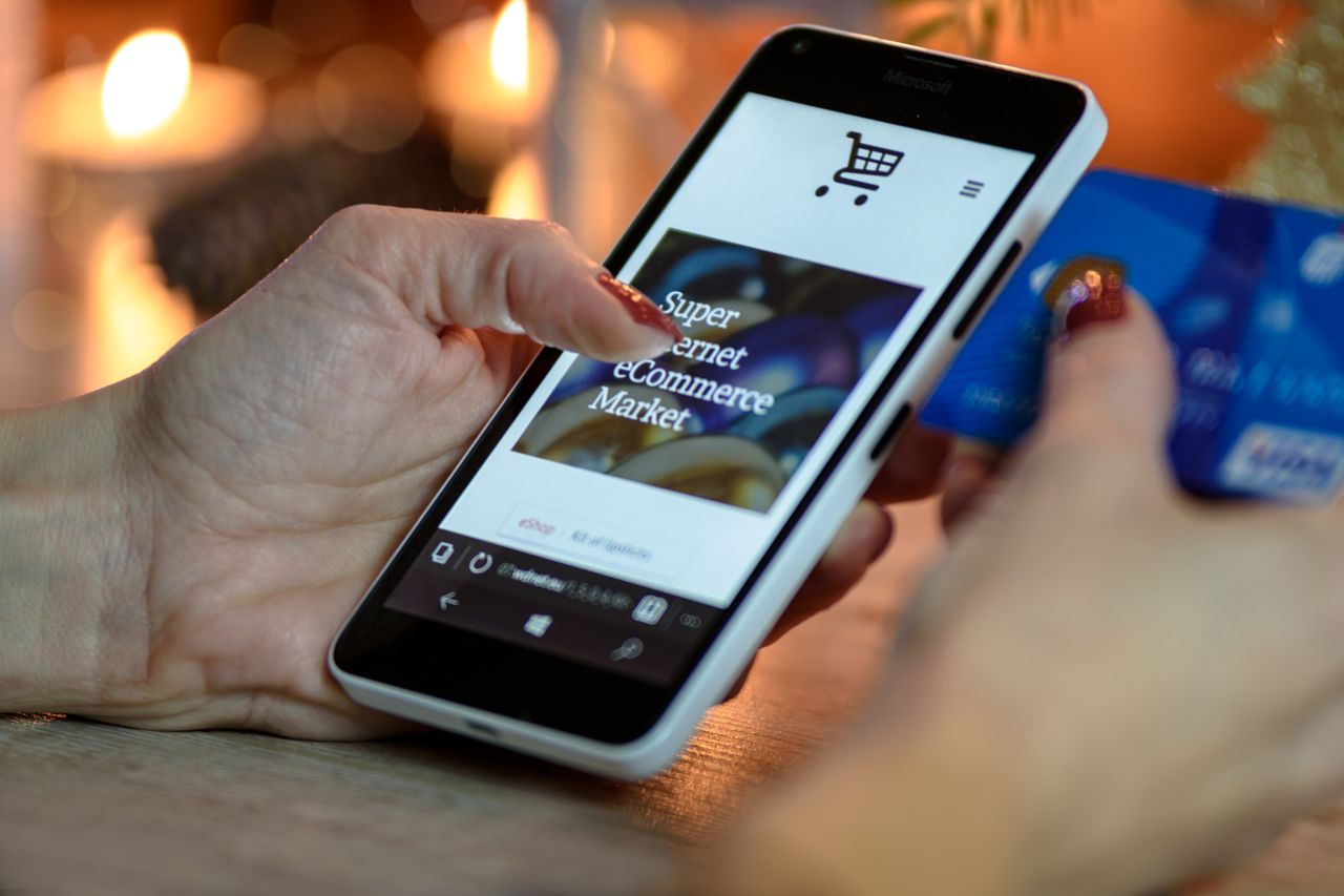 Femme utilisant un smartphone pour faire des achats en ligne, une carte de crédit à la main, avec un éclairage d’ambiance festif en arrière-plan.